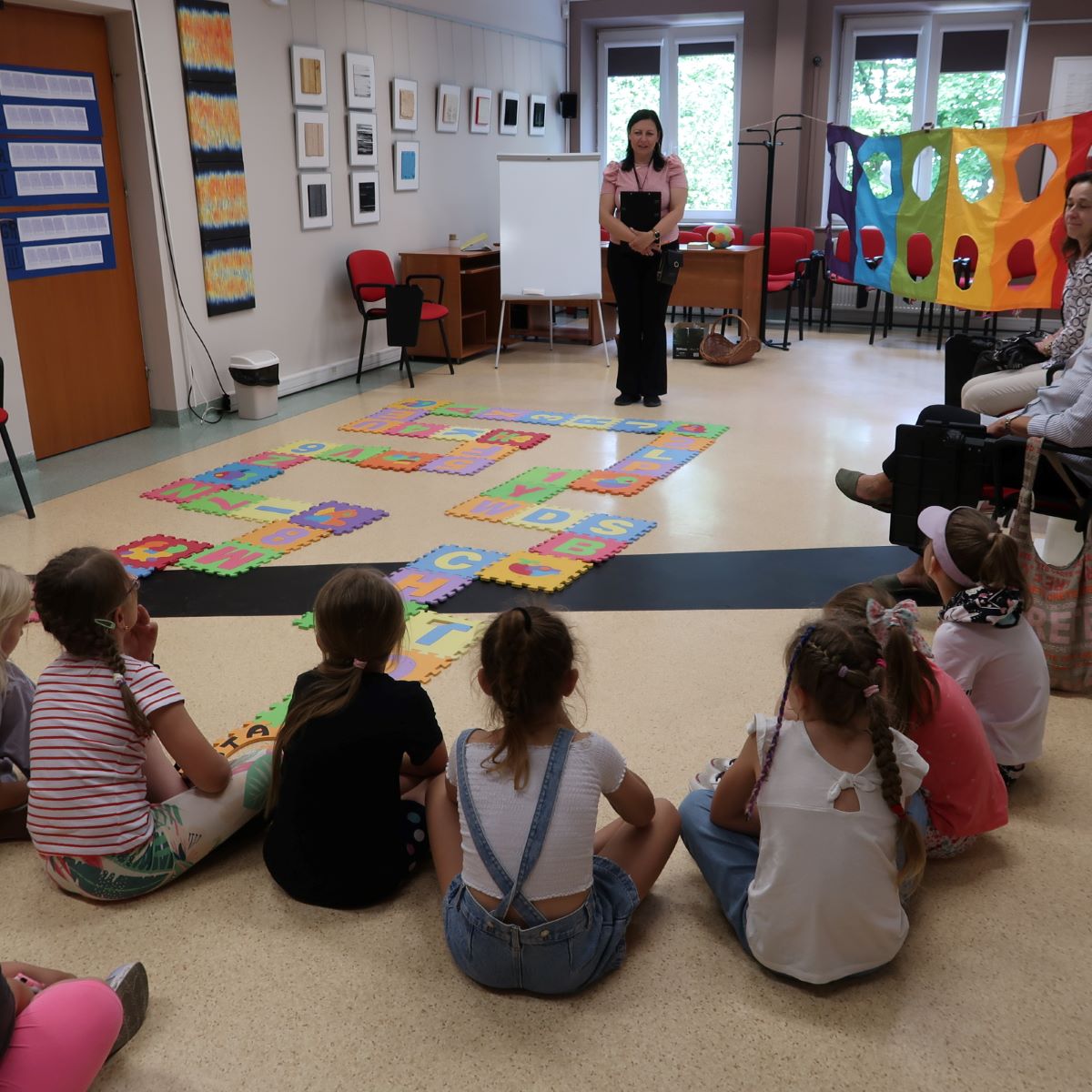 Dzieci siedząc na podłodze słuchają prowadzącej zajęcia pani.