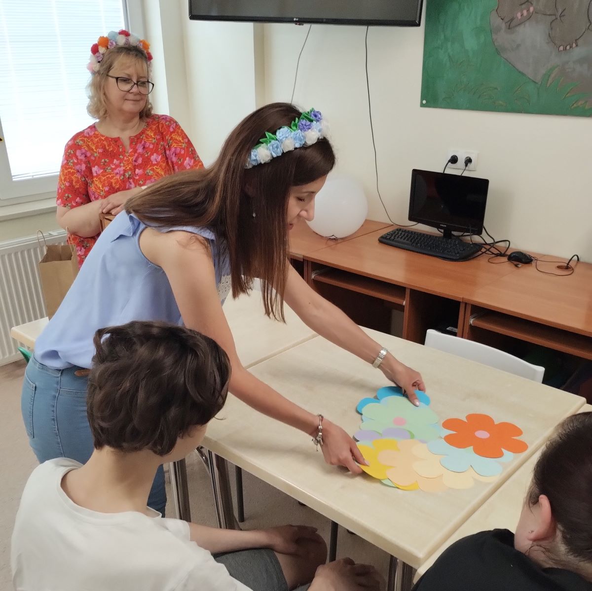 Uśmiechnięta kobieta z wiankiem na głowie stoi przy stole na którym rozłożone są papierowe wycinaki imitujące kolorowe kwiatki.