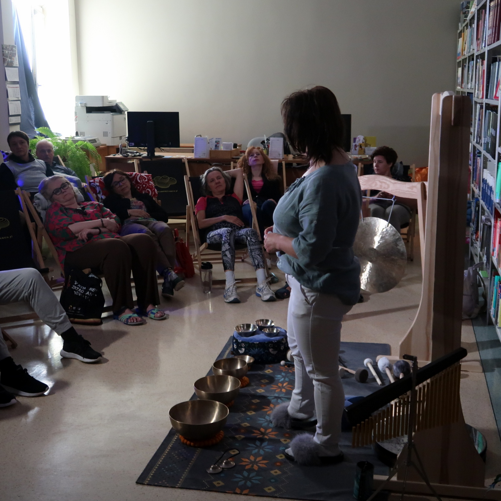 Uczestnicy warsztatów siedząc na leżakach słuchają kobiety stojącej w otoczeniu mis i talerzy