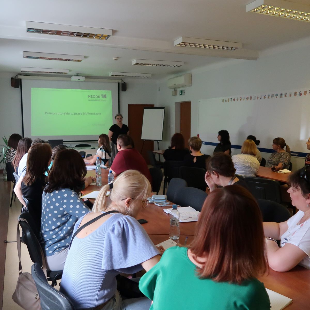 Grupa osób siedzi wokół stołów w sali lekcyjnej i uważnie słucha mówcy stojącego przed ekranem projektora.