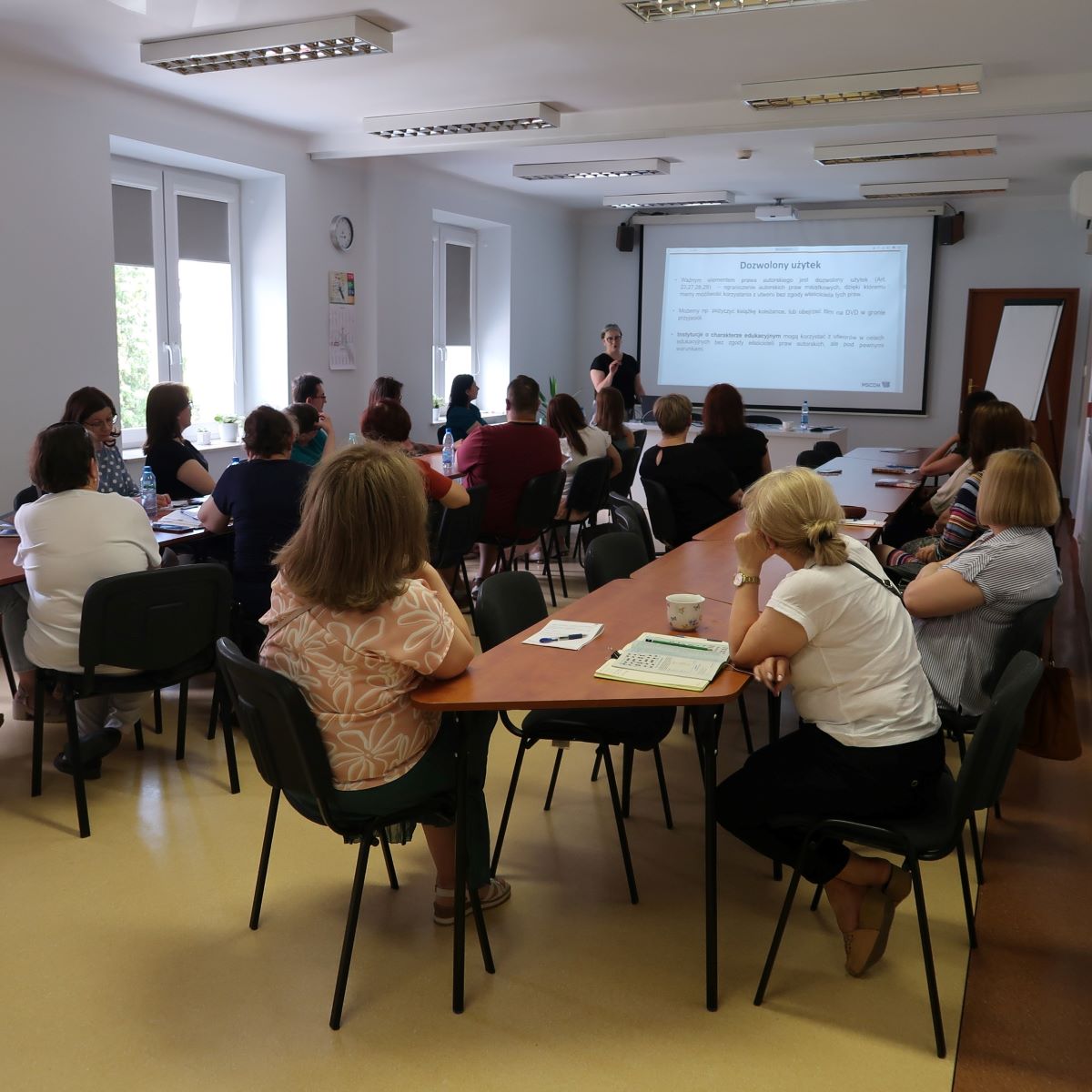 Grupa osób siedzących wokół dużego stołu konferencyjnego słucha prowadzącej stojącej przed ekranem wyświetlającym prezentację.