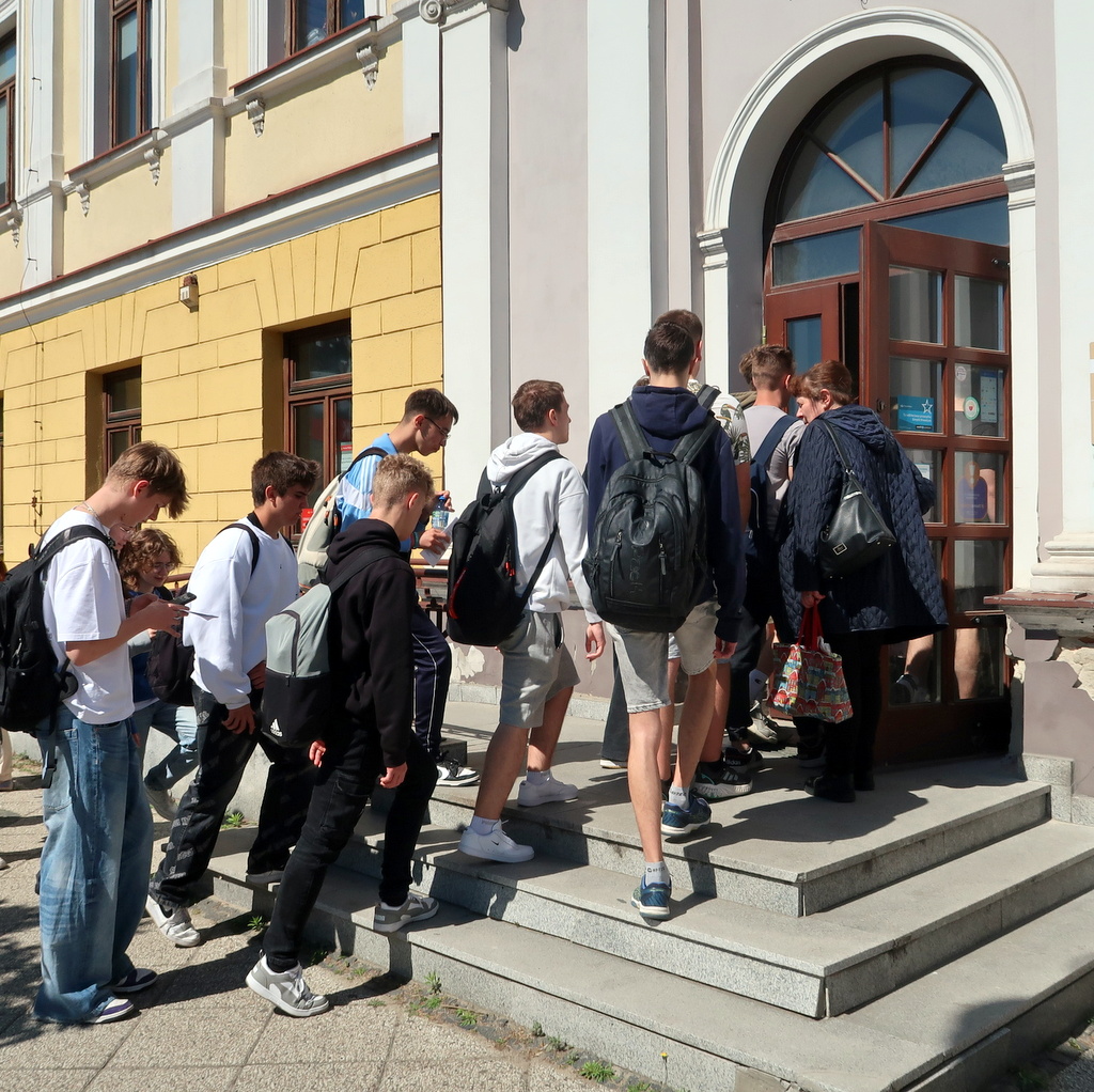 Grupa nastolatków wchodzi po schodach do budynku z dużymi łukowymi drzwiami.