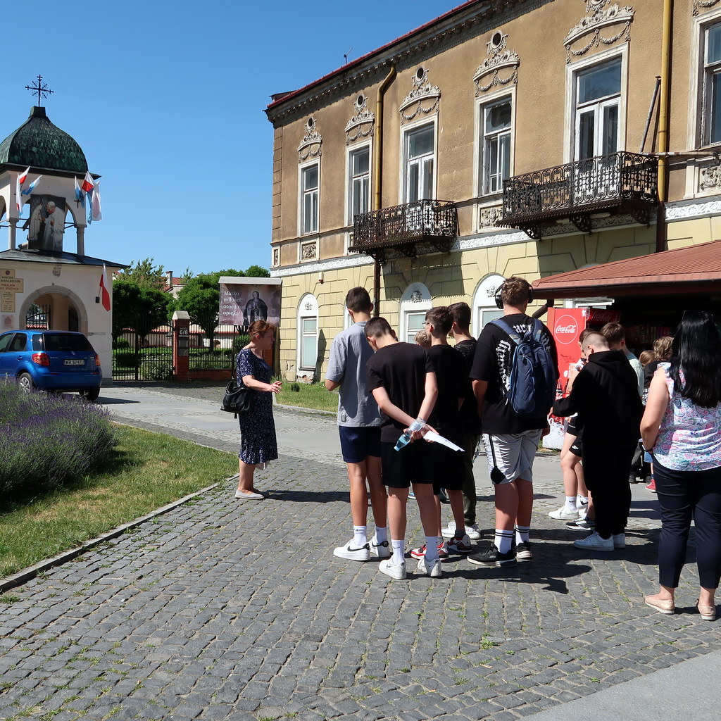 Grupa wycieczkowa zbiera się na brukowanej ścieżce przed zabytkowym budynkiem z ozdobnymi balkonami. Z grupą rozmawia przewodnik w niebieskiej sukience.
