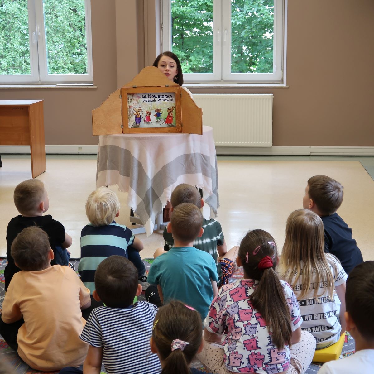 Młoda kobieta czyta dzieciom siedzącym na dywanie bajkę z teatrzyku kamishibai