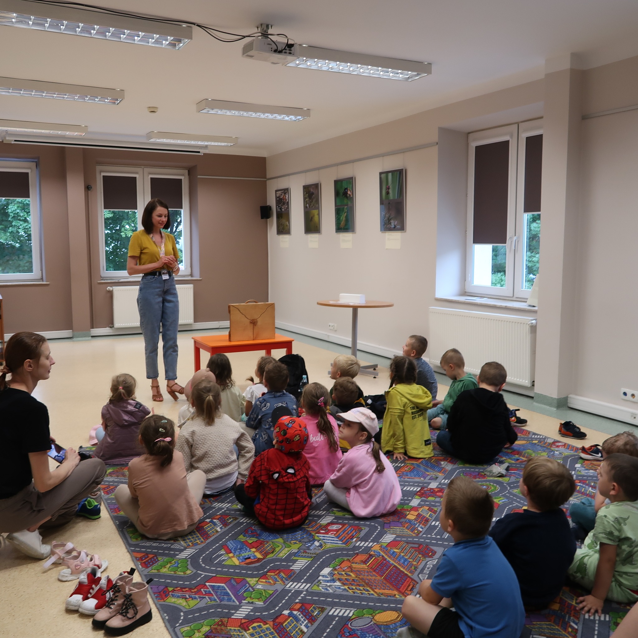 Dzieci siedząc na dywanie uważnie słuchają pani prowadzącej zajęcia.