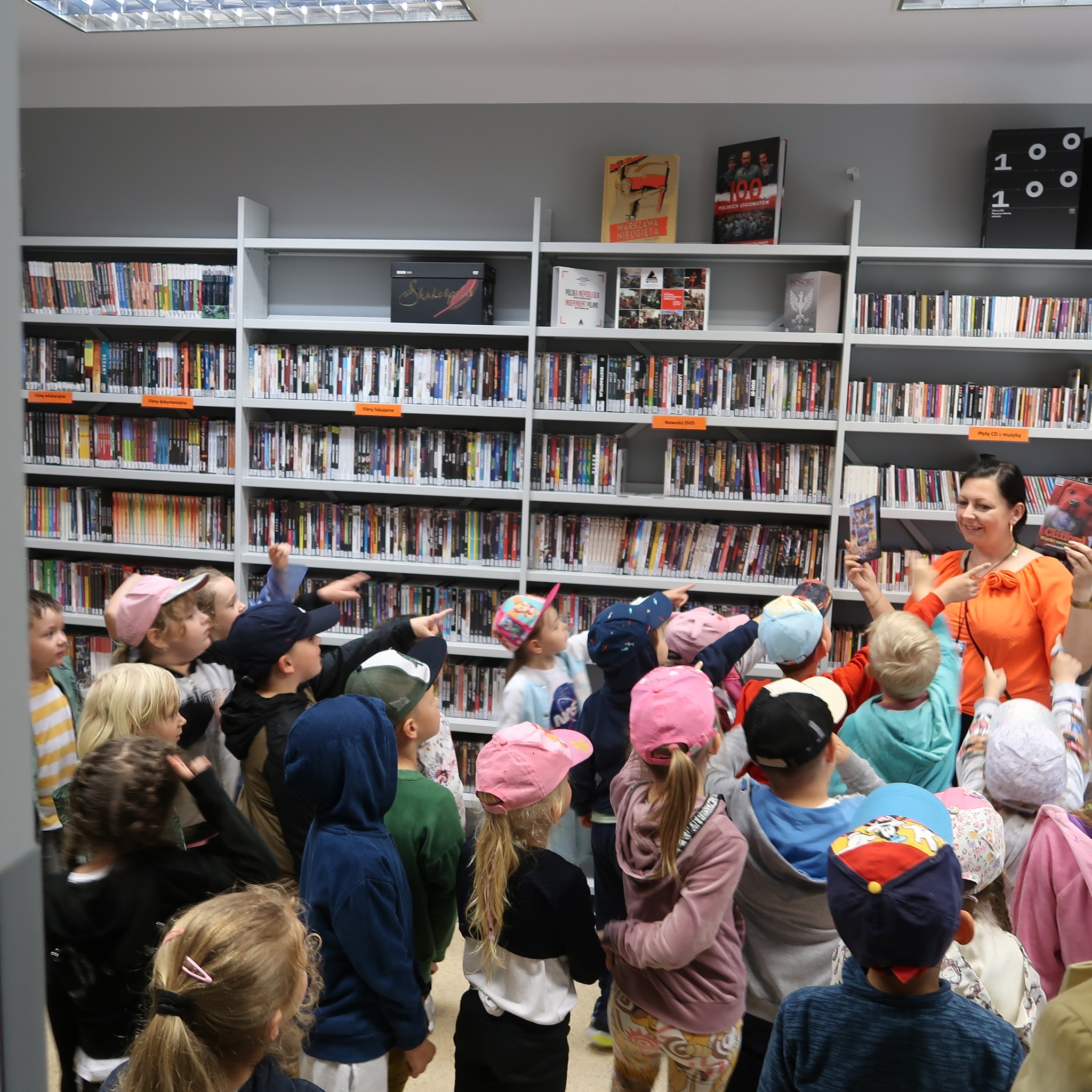 Grupa dzieci wskazuje palcami na okładki filmów video prezentowanymi przez panią