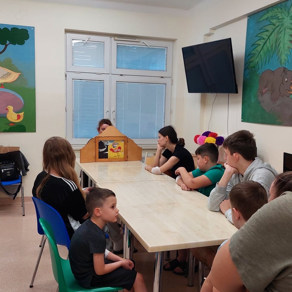 Zgromadzone na sali dzieci uważnie słuchają opowieści w formie teatrzyku kamishibai.