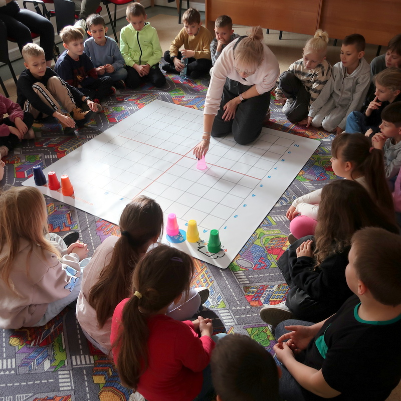 Dzieci siedząc na dywanie uważnie przyglądają się temu co pokazuje pani na planszy