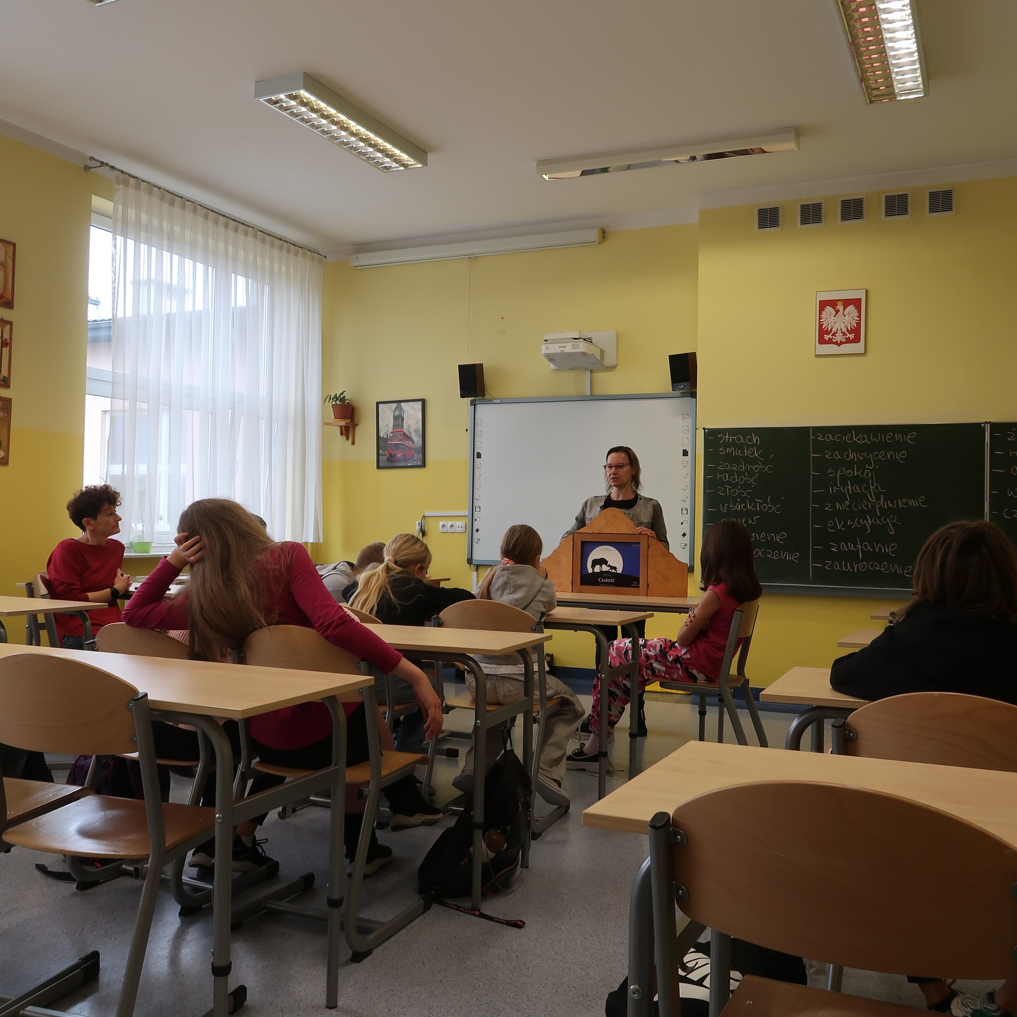 stylowa-zemsta Zajęcia w klasie szkolnej. Pani stojąc przodem do klasy prowadzi lekcję, prezentuje planszę z teatrzyku kamishibai.