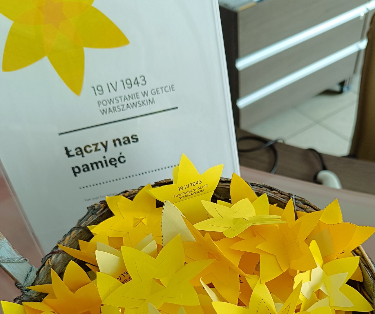 Kosz wypełniony żółtymi papierowymi żonkilami stoi na stole obok plakatu z napisem „19 IV 1943 Powstanie w getcie warszawskim” oraz hasłem „Łączy nas pamięć”. W tle widać fragment wnętrza z szufladami i krzesłem.