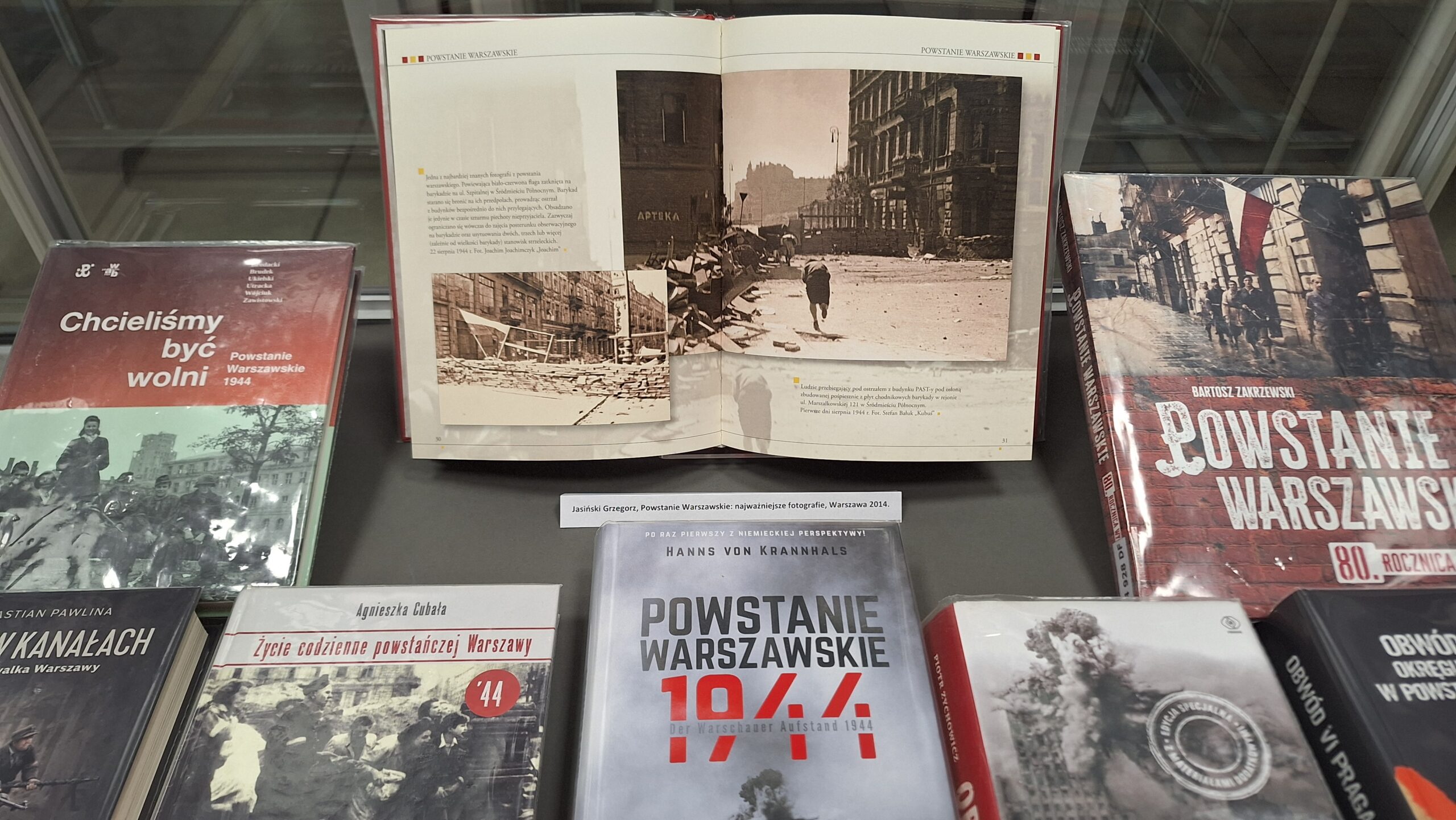 ystawa książek i albumów poświęconych Powstaniu Warszawskiemu zaprezentowana w szklanej gablocie. W centrum otwarta publikacja przedstawiająca archiwalne, czarno-białe fotografie zniszczonego miasta.